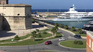 Una zona costera con una fortaleza histórica, un puerto deportivo con yates y una carretera con vehículos.