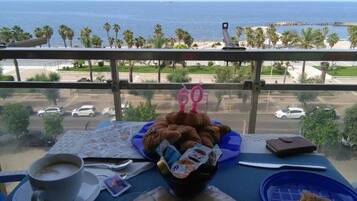 Een balkon met een ontbijttafel, een kop koffie, een schaal gebak en uitzicht op de oceaan en palmbomen.