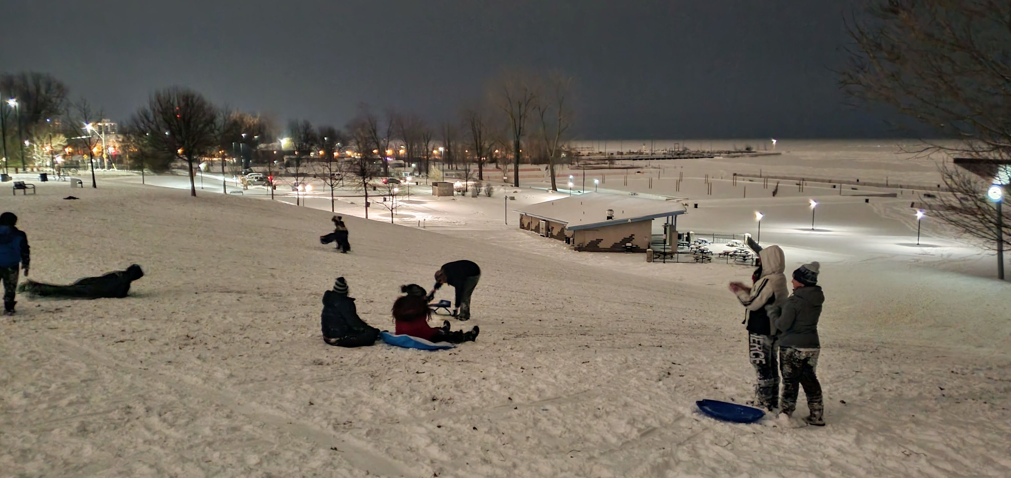 A snowy outdoor area with people sitting and standing, a small shelter, and streetlights.