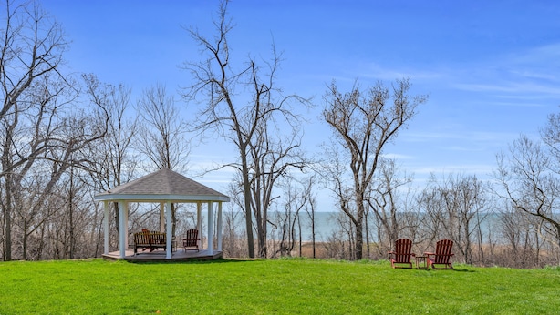 A gazebo with two Adirondack chairs in a grassy field, surrounded by leafless trees and a view of the water.