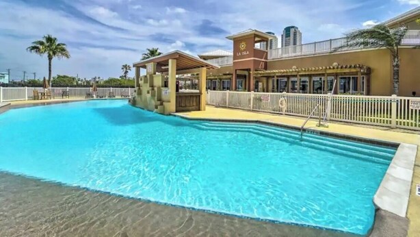 A swimming pool with a covered area, a building with a sign reading "LA ISLA", and a view of palm trees and a distant building. - Coral Cave Luxury Condo! Enjoy the beach, pools, BBQs, and bay views.  (South Padre Island)