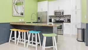 A kitchen with white cabinets, a granite countertop, a stainless steel refrigerator, a microwave, and a sink. There are three stools with colorful cushions. - Coral Cave Luxury Condo! Enjoy the beach, pools, BBQs, and bay views.  (South Padre Island)
