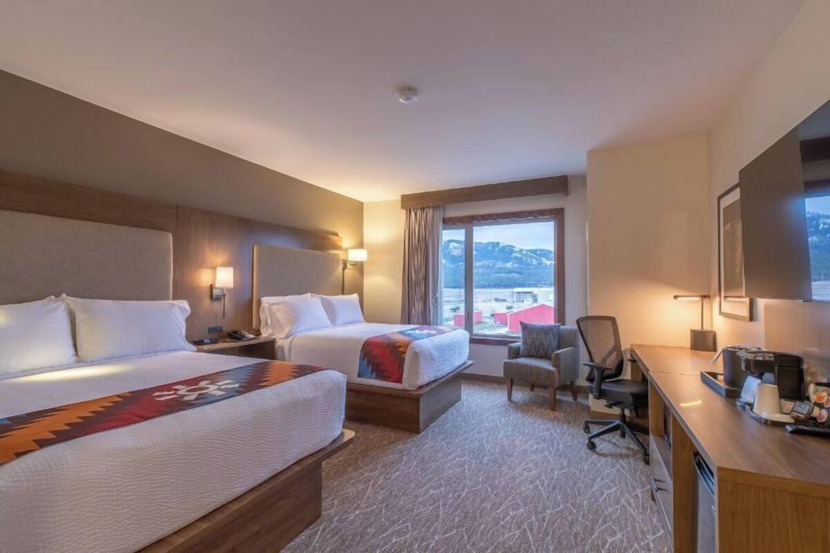 A hotel room with two beds, a desk, a chair, and a large window with a mountain view.