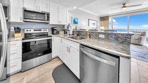 A modern kitchen with white cabinets, stainless steel appliances, a granite countertop, and a view of the ocean.
