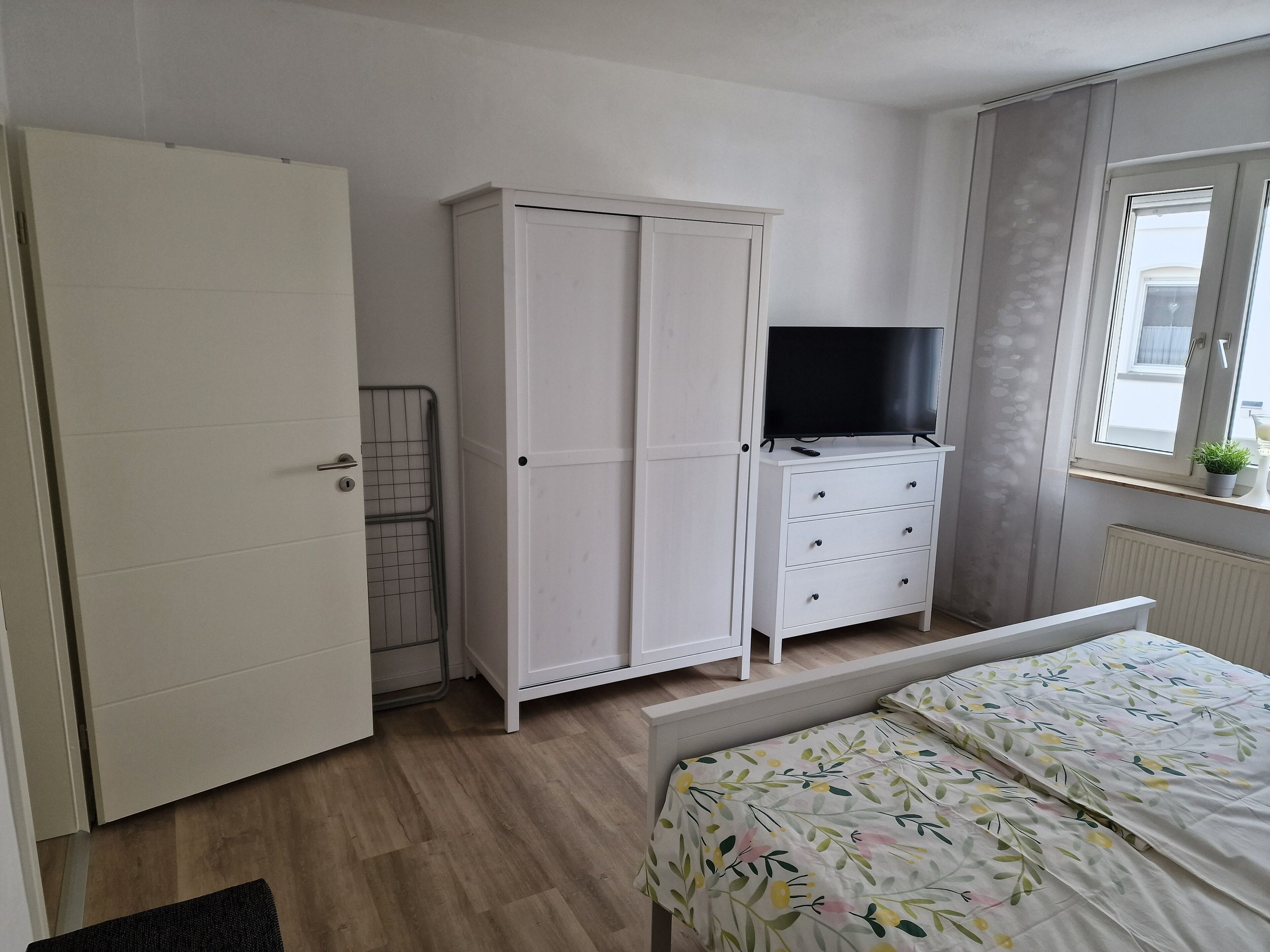 A bedroom with a bed, a wardrobe, a TV on a dresser, and a window with a plant.