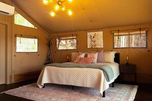 A bedroom with a bed, bedside tables, a chandelier, and windows. - NEW* “Wild Hare” Luxury Glamping Tent for 2 (Townsend)