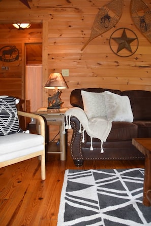 A cozy cabin interior with a wooden floor, a brown leather sofa, a white armchair, a patterned rug, and a lamp on a side table. - “Mystical Moose” Log Cabin (Townsend)