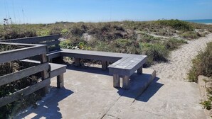 A concrete pathway leading to a wooden bench area with a picnic table, surrounded by beach vegetation and a clear view of the ocean.