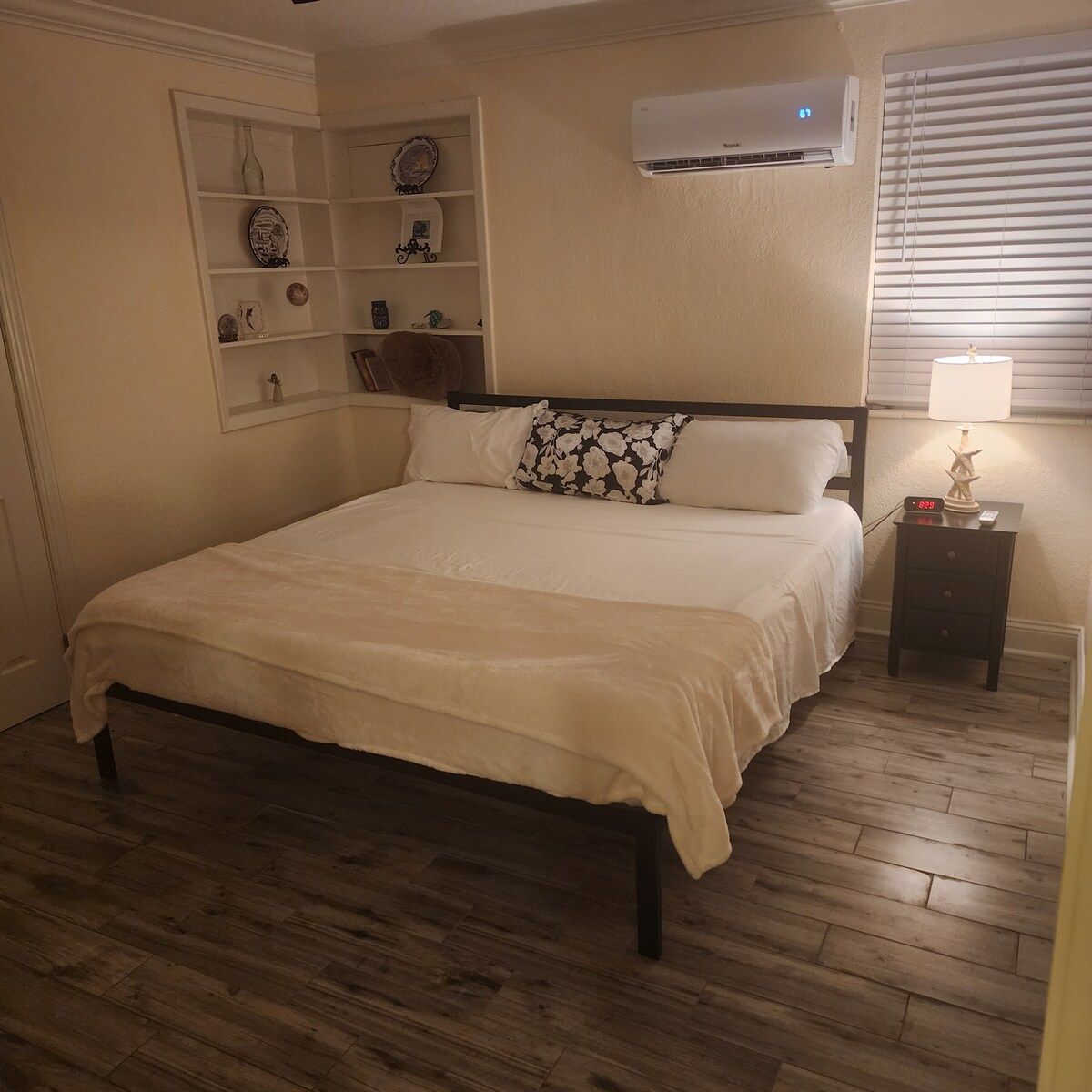 A bedroom with a bed, a nightstand, a wall-mounted shelf with decorative items, and an air conditioning unit.