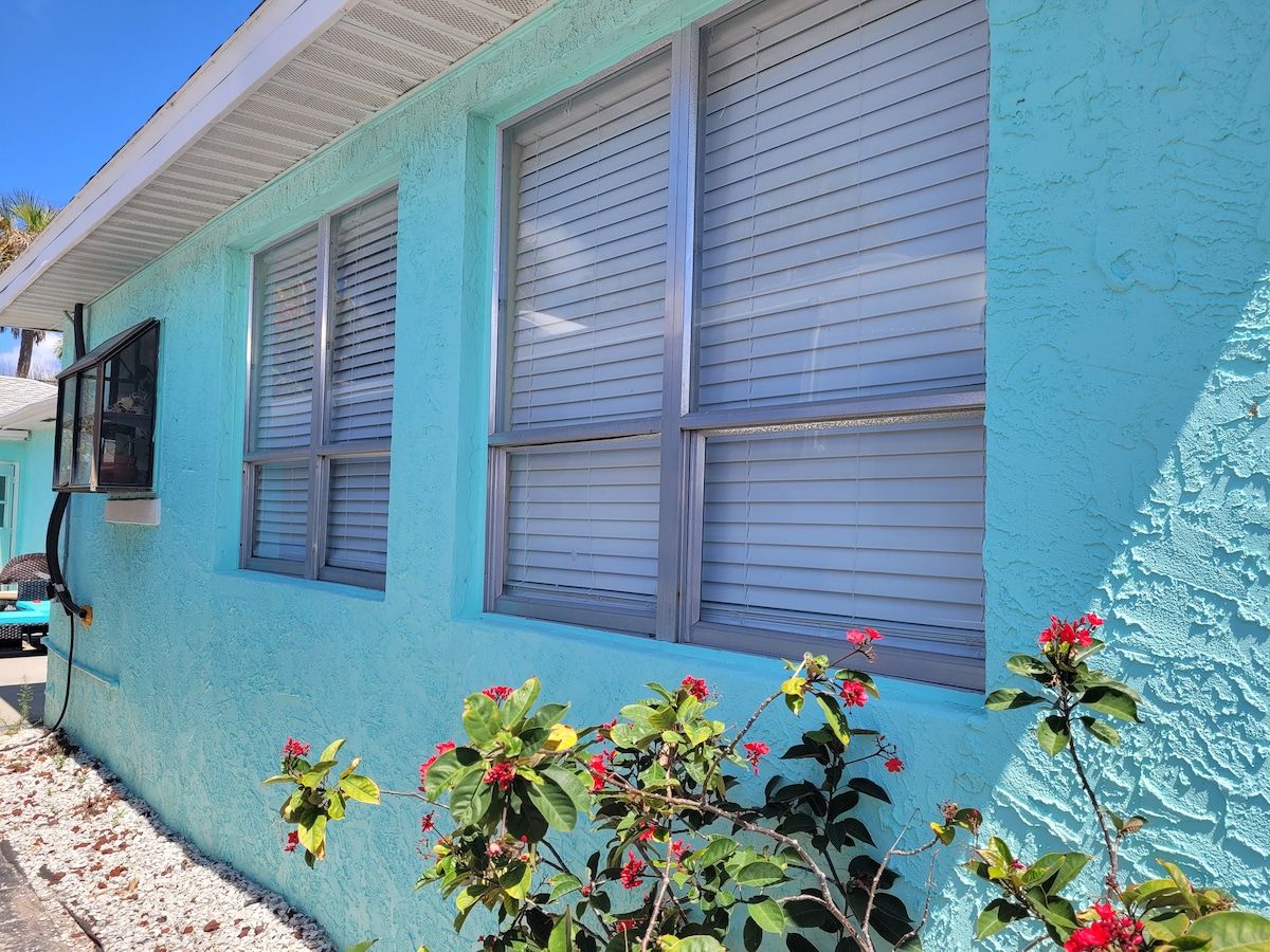 A building with a light blue exterior, two windows with closed blinds, and a small plant with red flowers in front.