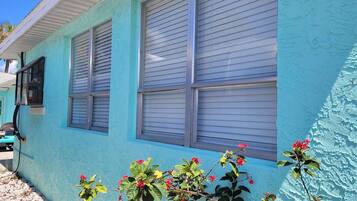 A building with a light blue exterior, two windows with closed blinds, and a small plant with red flowers in front.