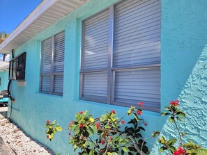 Un bâtiment à la façade bleu clair, doté de deux fenêtres dont les stores sont fermés, et d’une petite plante aux fleurs rouges placée devant.