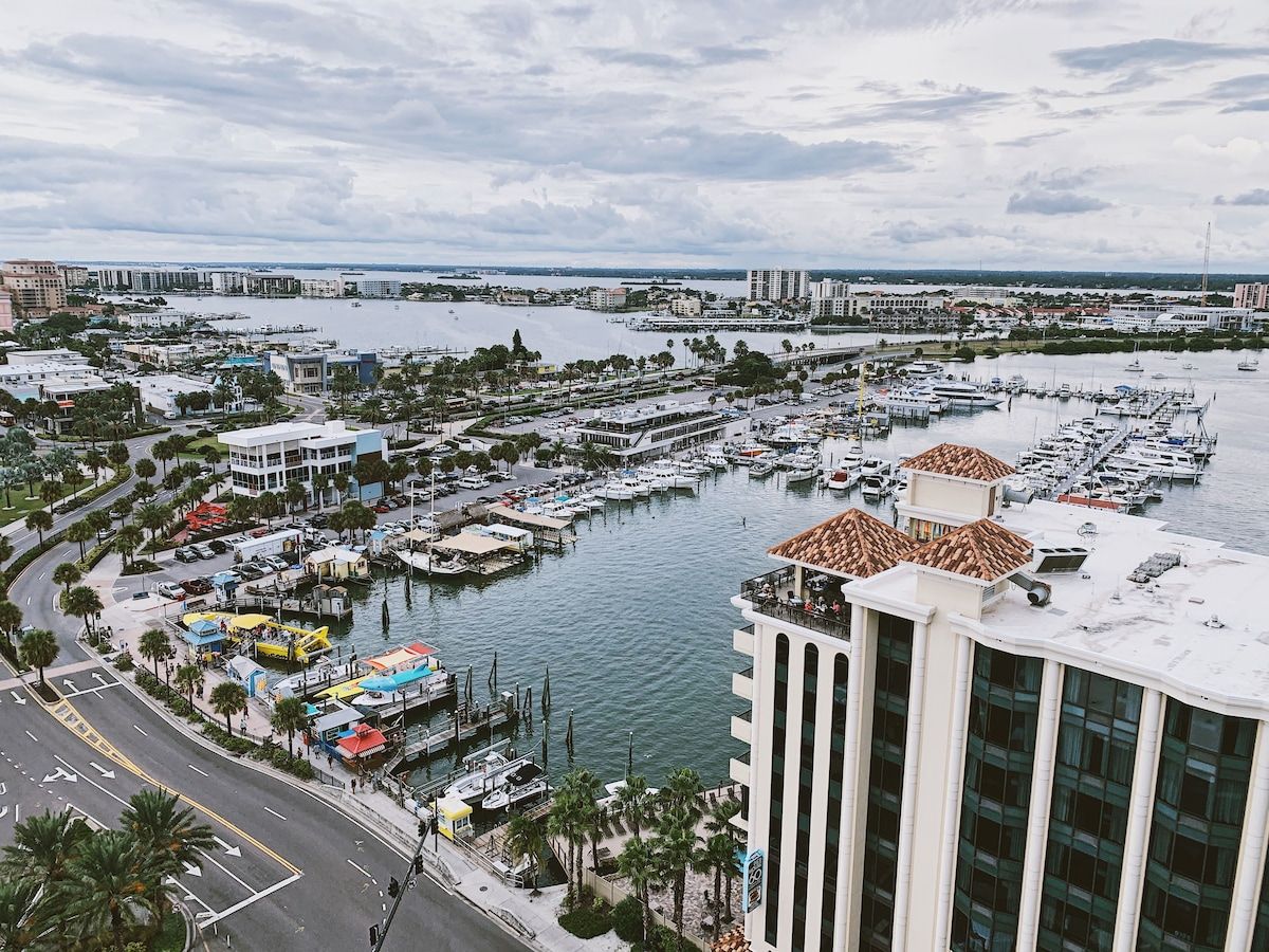 A marina with boats docked, a road with palm trees, and a large building with a white facade.