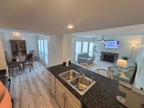A modern kitchen with a double sink, dining area with chairs, a sofa, and a TV displaying 'Saturday America'.