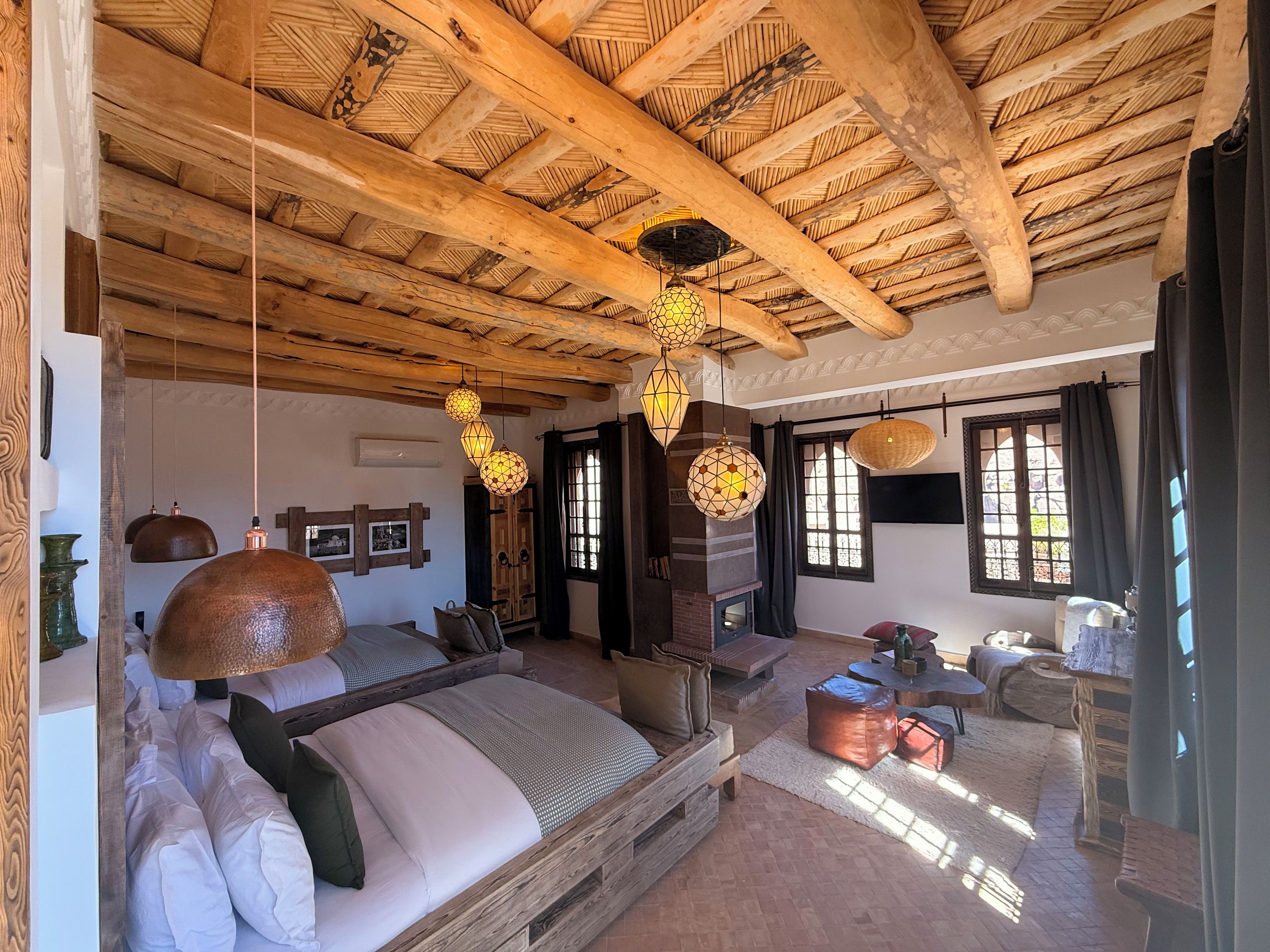 A rustic room with a wooden ceiling, a large bed, a fireplace, and hanging lamps.