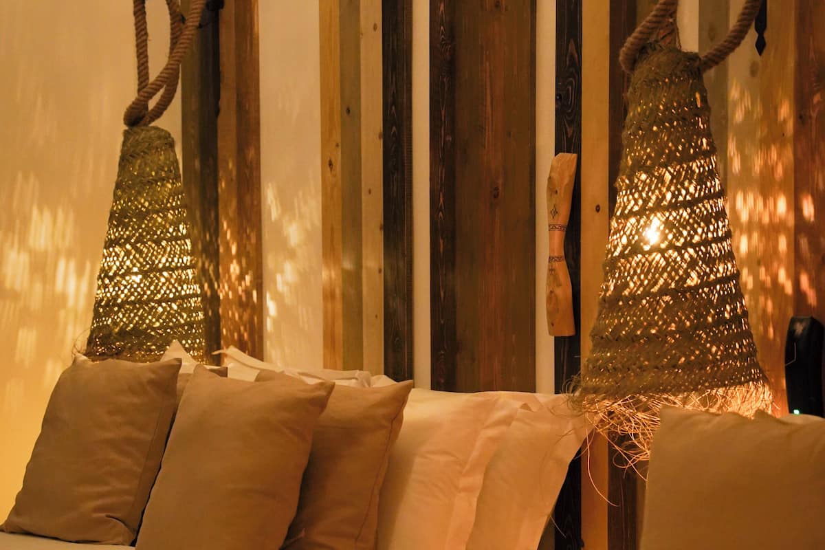 A bed with beige bedding, a wooden headboard, and two hanging lamps with a woven design.
