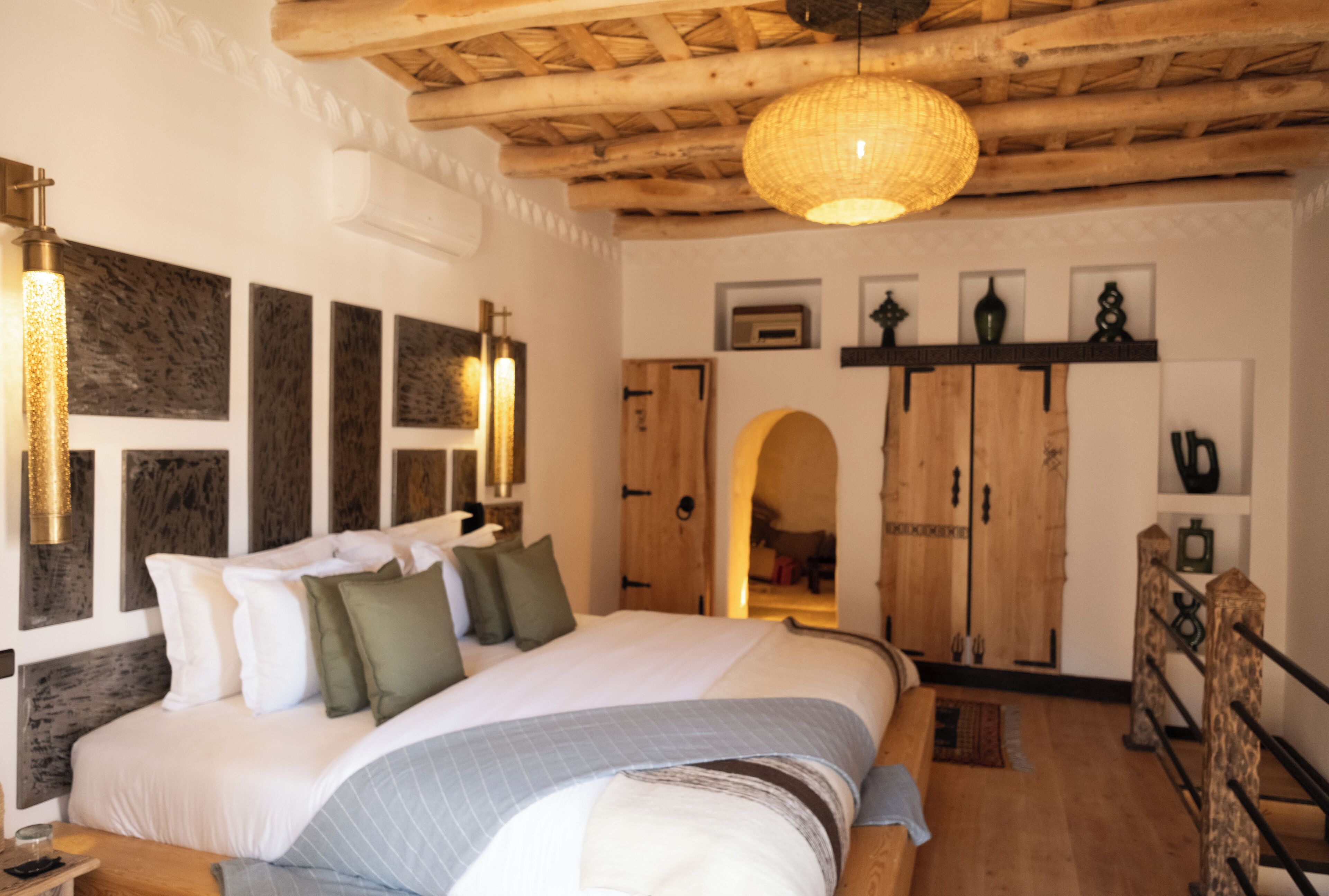 A bedroom with a large bed, wooden ceiling beams, a hanging light fixture, and a wardrobe with a mirror.