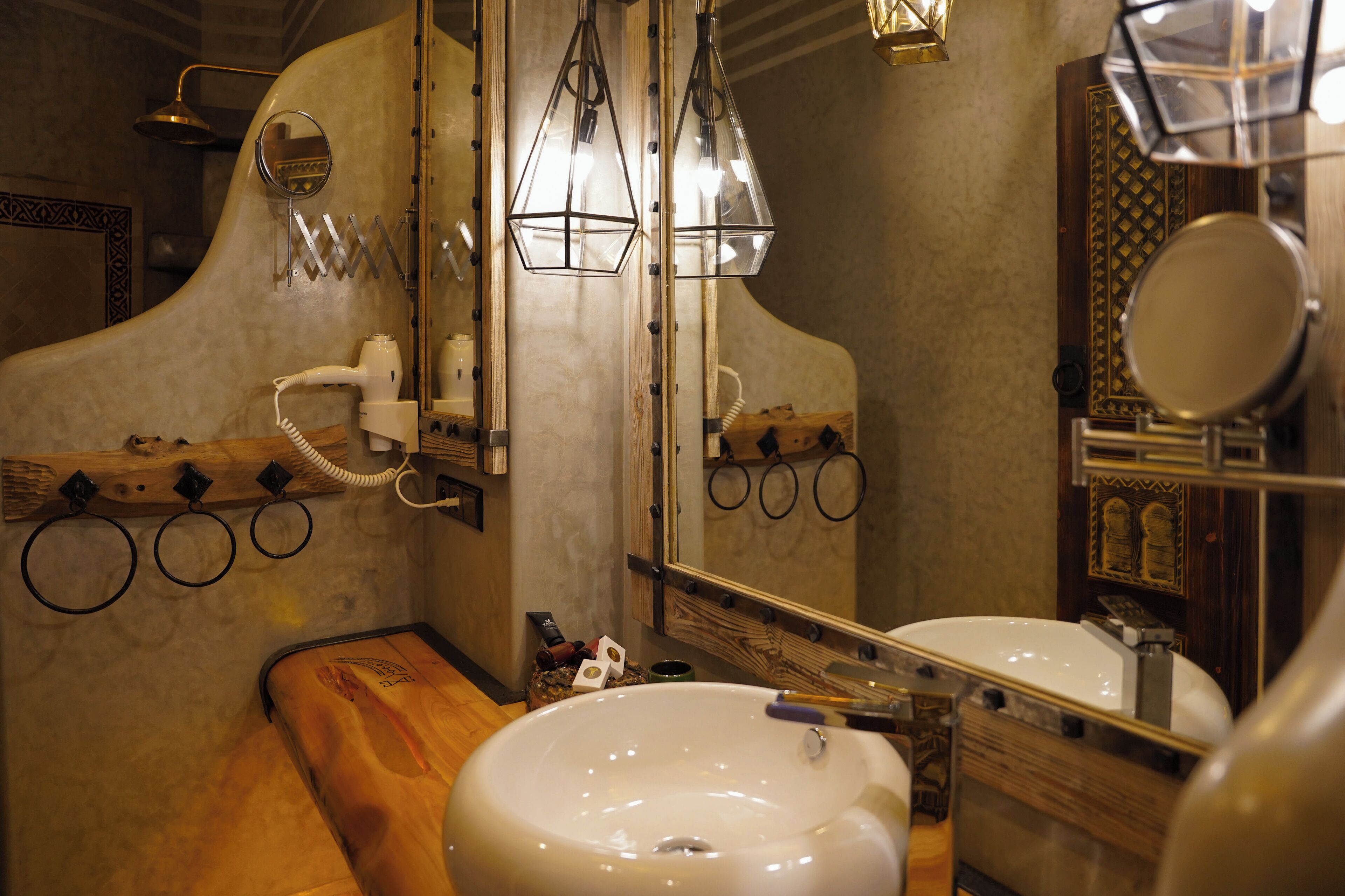 A bathroom with a wooden vanity, a white sink, and a mirror. There are two pendant lights, a towel rack, and a showerhead.