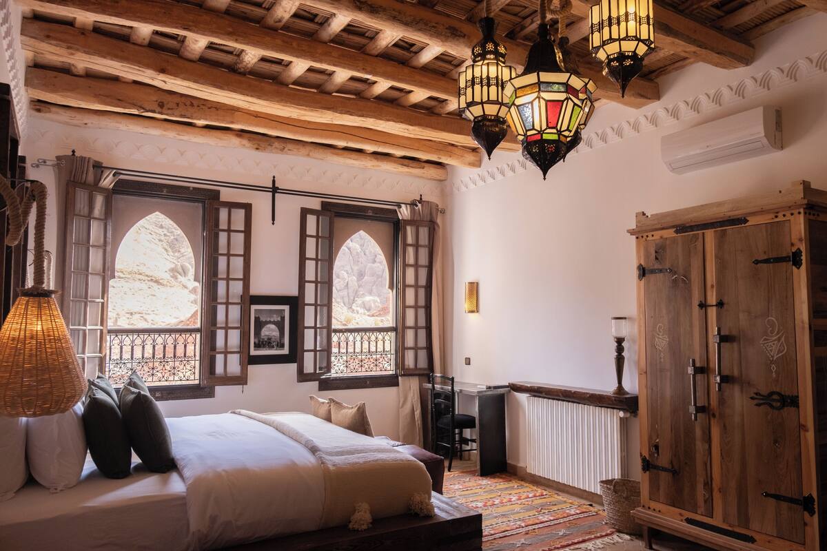 A traditional room with a bed, wooden ceiling beams, a large wardrobe, and a window with a view.