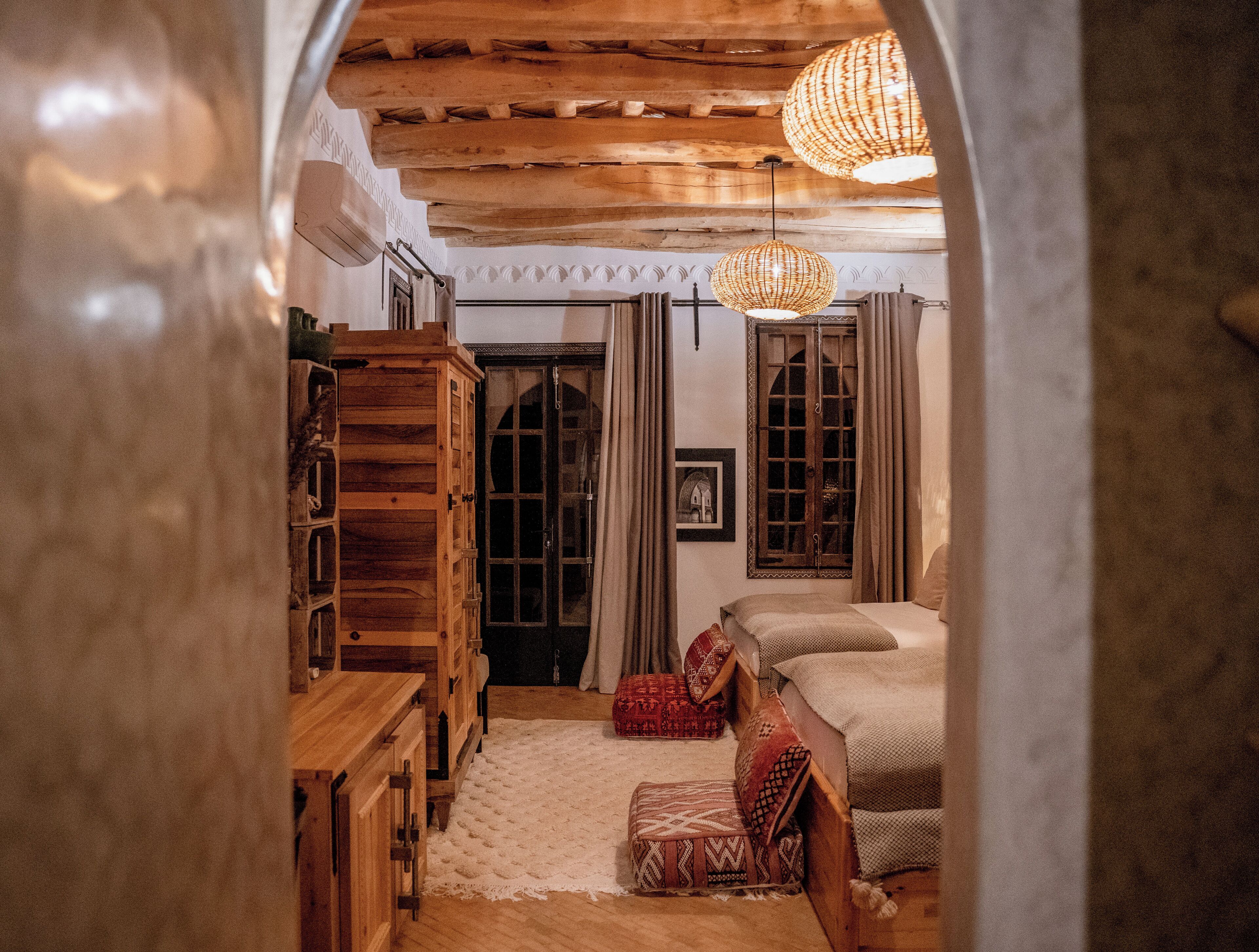 A cozy room with a wooden ceiling, a bed with patterned bedding, a wooden wardrobe, and a large window with curtains.