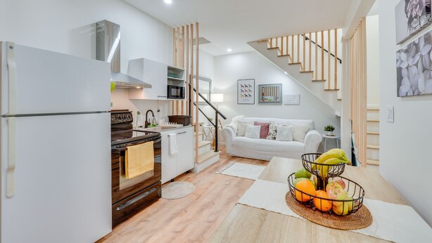 Cocina moderna con estufa, horno y refrigerador blanco. Se observa un comedor con una mesa de madera y una cesta de frutas. Una escalera conduce a una sala de estar con un sofá blanco y un reloj en la pared.