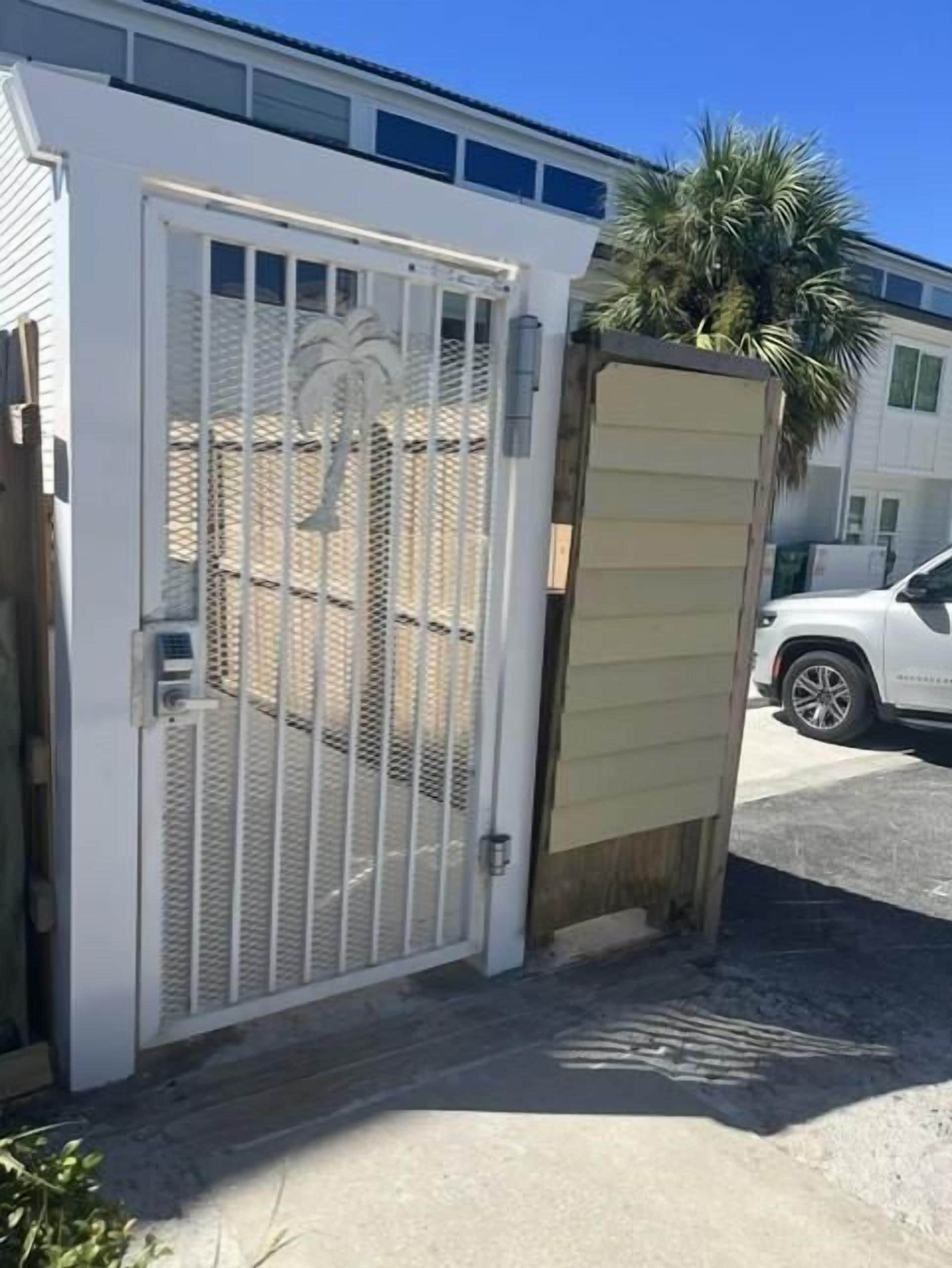 A gated entrance with a metal gate and a wooden door, a palm tree, and a white vehicle parked in the background.