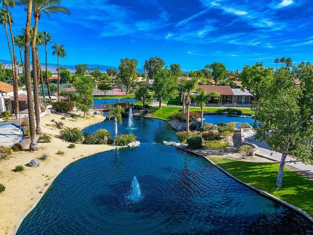 A swimming pool with a fountain, surrounded by palm trees and a view of houses in the background.