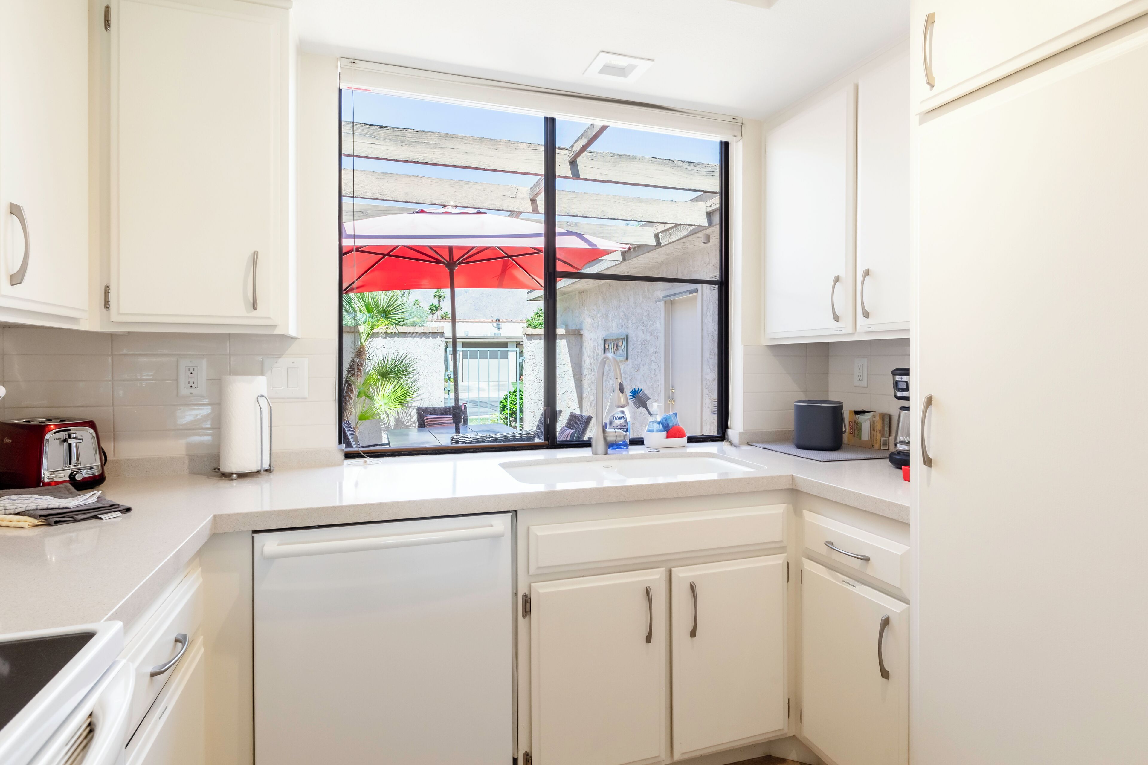 A kitchen with white cabinets, a window with a red umbrella, and a countertop with various items.