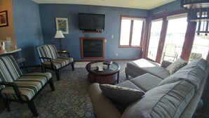 A living room with a sofa, striped chairs, a round coffee table, a TV, and a fireplace. - Snug Harbor Inn - Studio Suite 201 & 204 (Sturgeon Bay)