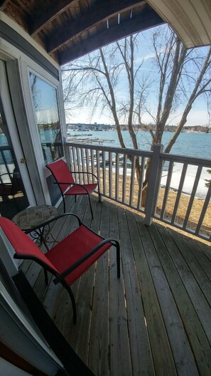 A wooden deck with two red chairs and a table, overlooking a body of water with boats and a tree. - Snug Harbor Inn - Studio Suite 201 & 204 (Sturgeon Bay)