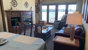 A room with a dining table, chairs, a sofa, a lamp, and a view of a body of water through large windows. - Snug Harbor Inn - Suite 202 & 203 (Sturgeon Bay)