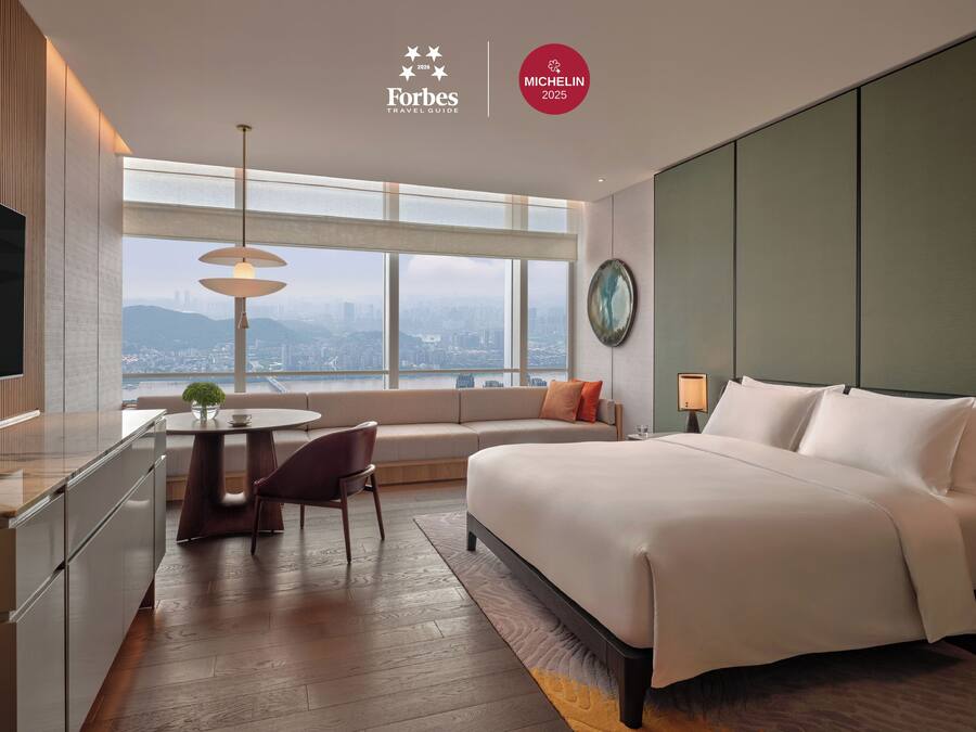 A modern hotel room with a large bed, a dining area, and a view of the city.