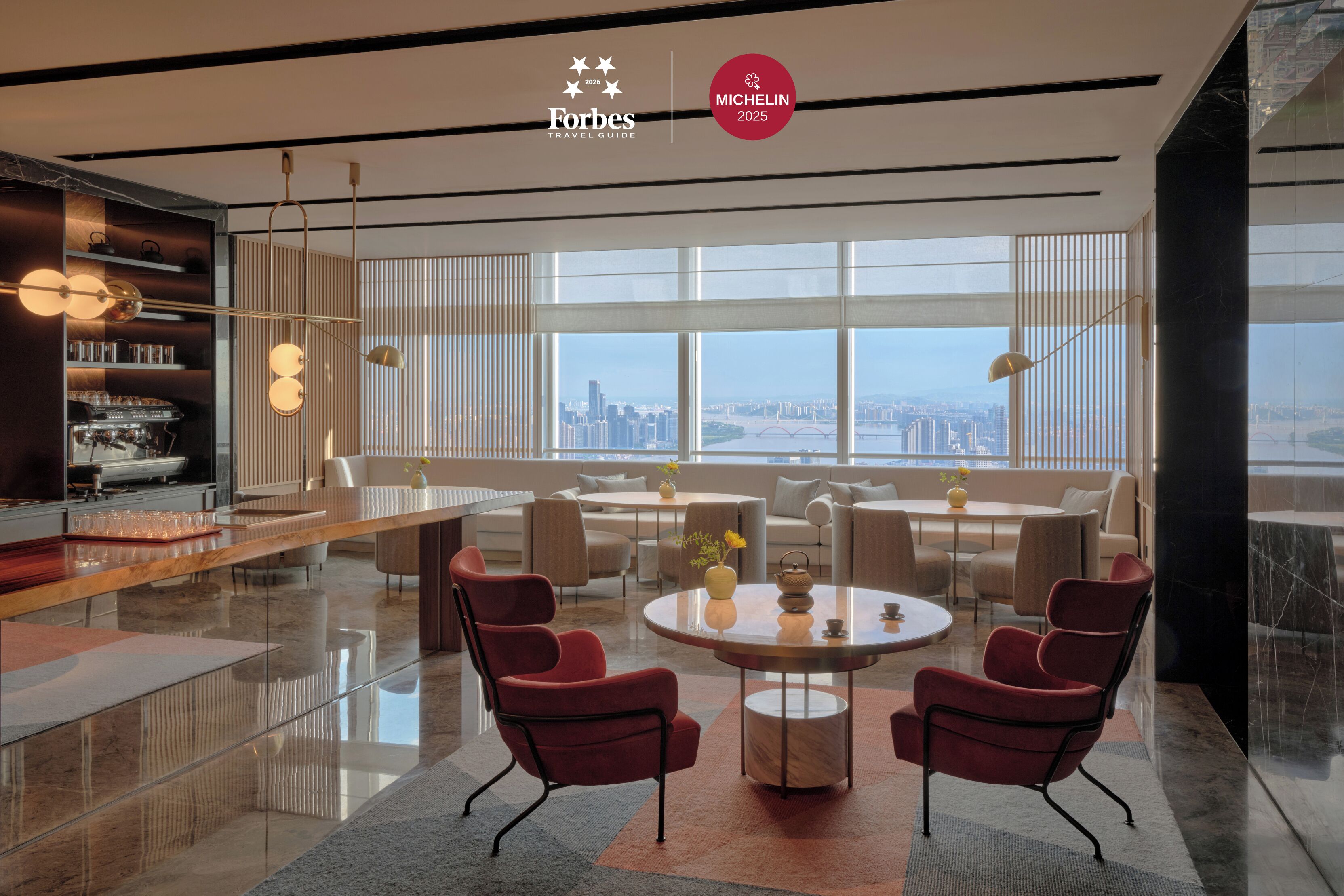 A modern hotel lobby with a large round table, red chairs, a bar area, and large windows offering a city view.