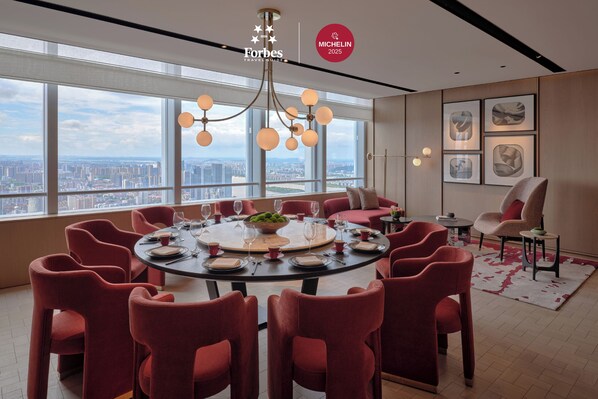 Habitación de hotel en un rascacielos con una gran mesa redonda dispuesta para comer, sillas tapizadas en rojo, vistas a la ciudad y decoración moderna.