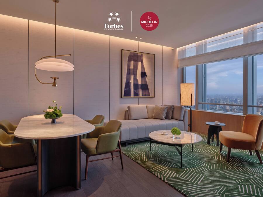 A modern hotel room with a dining area, a sofa, and a large window with a city view.