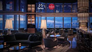 A modern lounge area with seating, a bar, and large windows offering city views.