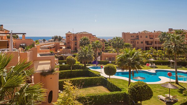 A resort with a swimming pool surrounded by palm trees and sun loungers, with buildings in the background.