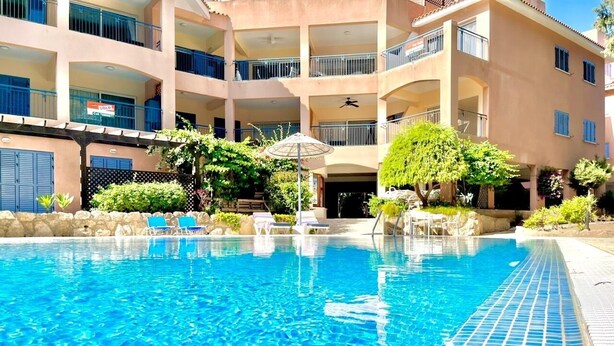 A swimming pool with lounge chairs and a covered area, surrounded by a multi-story building with balconies. - Beautifully presented 2 bedrooms apartment with a pool and garden view. (Paphos)