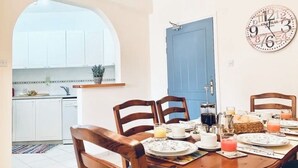 A dining area with a wooden table set for four, surrounded by wooden chairs. A blue door is visible in the background, and a wall clock is mounted above the table. - Beautifully presented 2 bedrooms apartment with a pool and garden view. (Paphos)