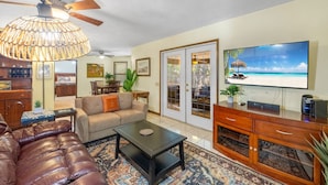 A living room with a sofa, coffee table, and TV on a wooden cabinet. There is a ceiling fan and a large window with a view of the ocean. - Enjoy art and relaxation in this hilltop home (Cedar Key)