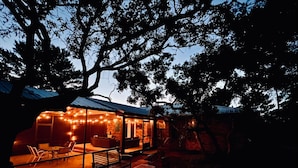 A cozy outdoor seating area with a glass enclosure, surrounded by trees and featuring string lights. - Enjoy art and relaxation in this hilltop home (Cedar Key)