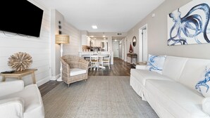 A modern living room with a white sofa, a wicker chair, a flat-screen TV mounted on the wall, and a dining area with a white table and chairs. - Ocean Breeze East 803 (Pensacola)