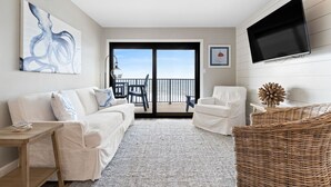 A modern living room with a white sofa, a white armchair, a flat-screen TV, and a large window with a view of the sea. - Ocean Breeze East 803 (Pensacola)