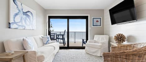 A modern living room with a white sofa, a white armchair, a flat-screen TV, and a large window with a view of the sea.