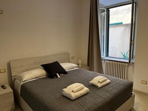 Un lit bien fait, recouvert d’une couverture sombre, avec deux oreillers blancs et un oreiller décoratif noir. Deux serviettes pliées sont posées sur le lit. La chambre est dotée d’une tête de lit en bois, d’une table de chevet avec une lampe et d’une fenêtre avec des rideaux.
