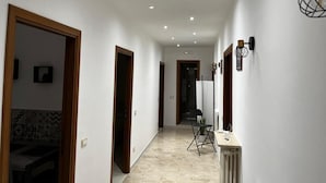 Un couloir aux murs blancs, avec des portes en bois, des spots encastrés au plafond, un radiateur et une chaise agrémentée d’une plante.