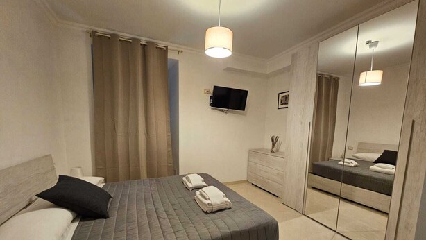 Een moderne hotelkamer met een groot bed, een aan de muur bevestigde televisie, een dressoir en een spiegel die het bed weerspiegelt.