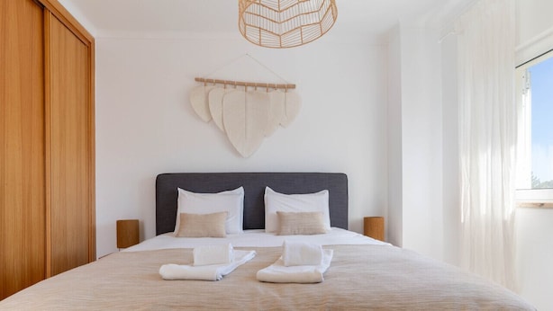 A neatly made bed with a beige bedspread, two pillows, and two towels. There is a wooden wardrobe to the left and a wall-mounted light fixture above the bed. - Vivenda Duneta - in Walking Distance to the Beach by Lisbeyond (Aljezur)