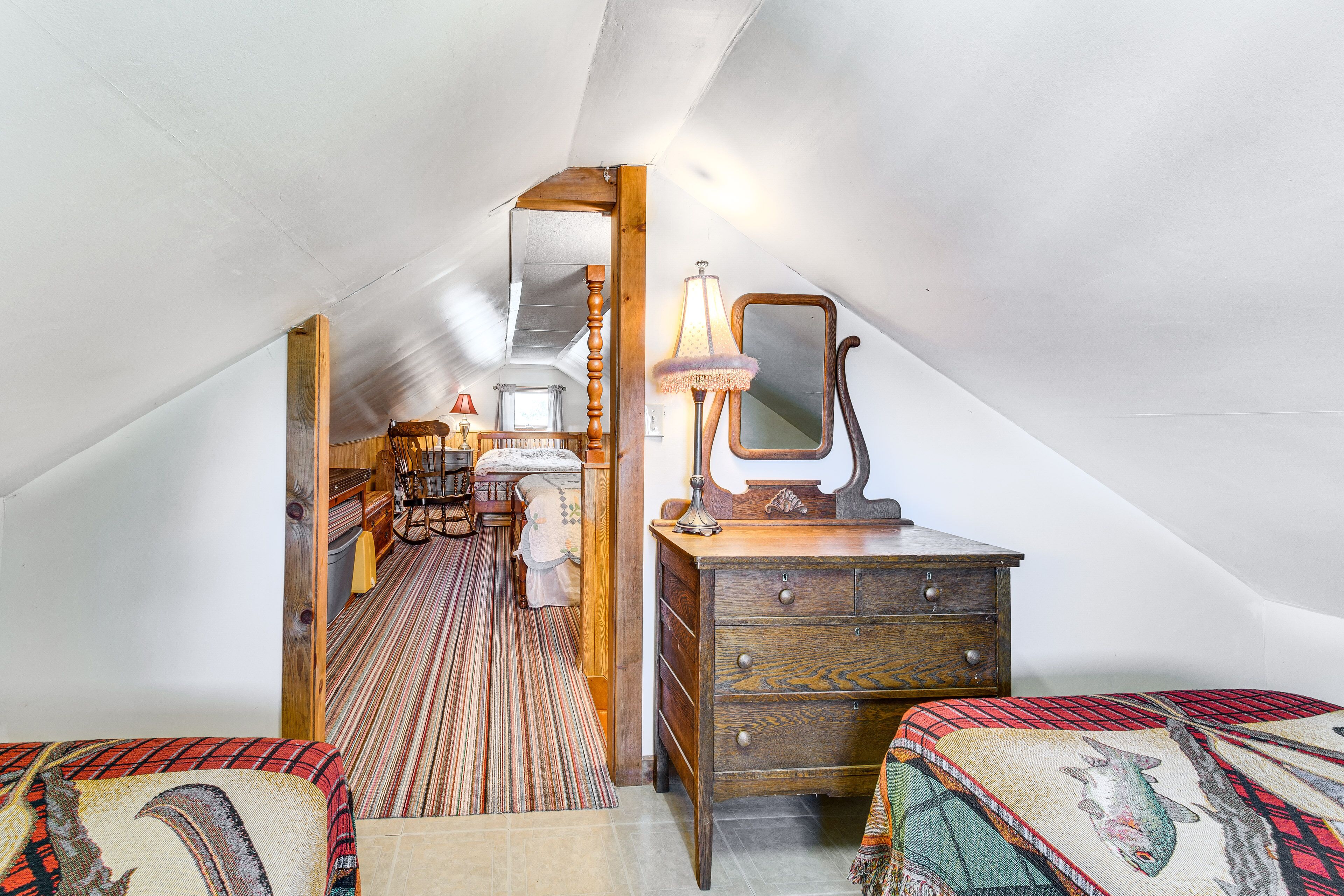 Een gezellig zolderkamer met een houten dressoir, twee bedden met beddengoed met een patroon, een spiegel en een lamp.