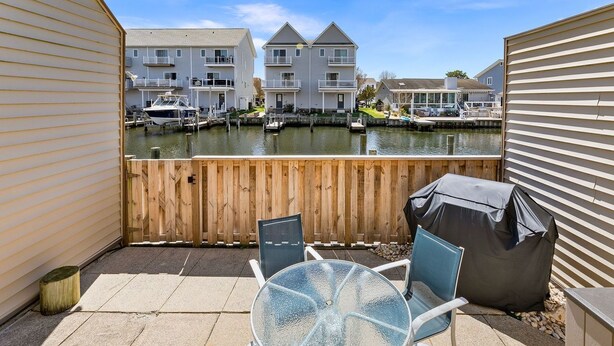 Eine Terrasse mit Glastisch und Stühlen, einem überdachten Grill und Blick auf einen Yachthafen mit Booten und Gebäuden im Hintergrund.